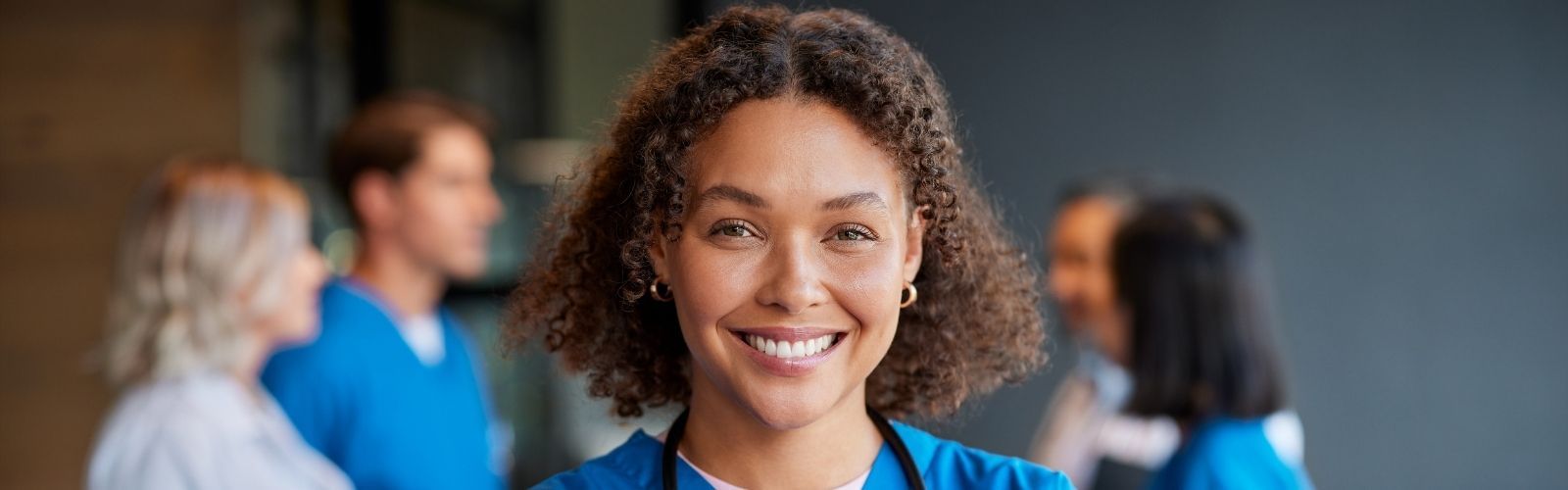 A female nurse smiling at the camera with a group of doctors and nurses consulting each other behind.